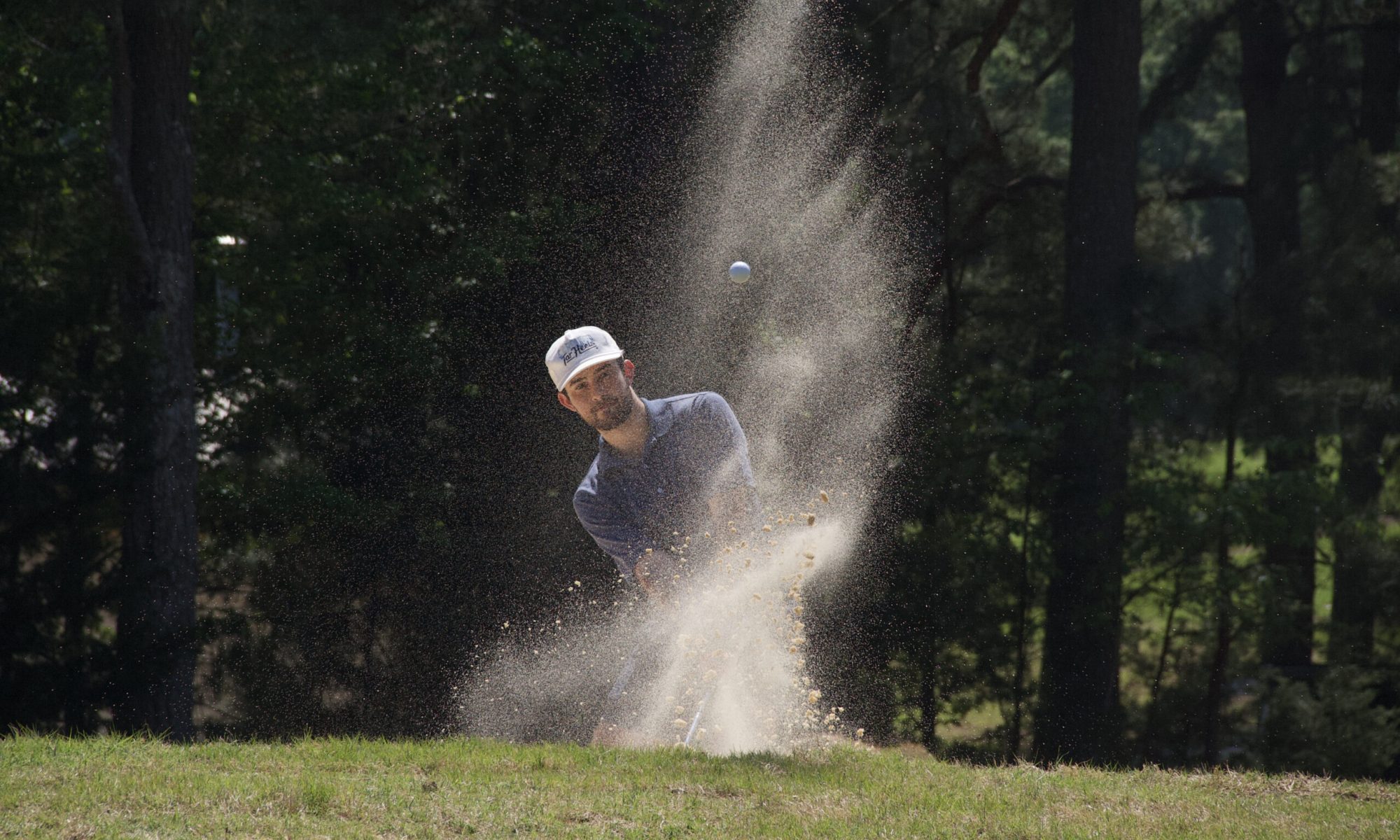 an image of a amateur golfer hitting a bunker shot from just off the green. sand and the ball are seen flying in the air.